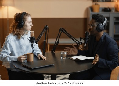 ポッドキャスト中にスーツを着た黒人男性を身振りするために話しているカップと白人の女性のサイドビュー，両方のスタジオで丸いテーブルに座っている間、 ヘッドフォンを着て写真素材2419303977Shutterstock